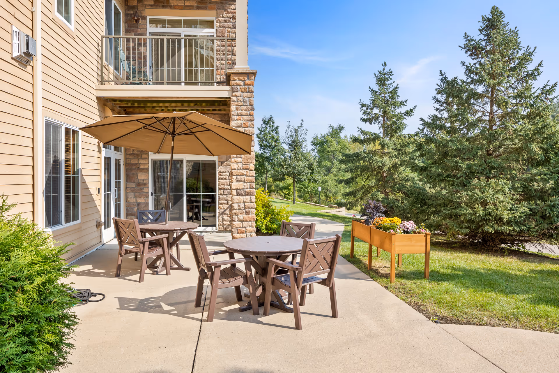 Outdoor patio area at Founders Ridge with two round tables, each surrounded by four chairs. One table has a large beige umbrella providing shade. The patio is adjacent to a building with beige siding and stone accents. There are green shrubs and trees nearby, along with a raised wooden planter box containing colorful flowers. The sky is clear and blue.