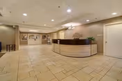 Spacious reception area with a curved wooden front desk, beige tiled floor, recessed ceiling lights, and neutral-colored walls. There is a decorative wall piece behind the desk and a door to the right side of the desk.
