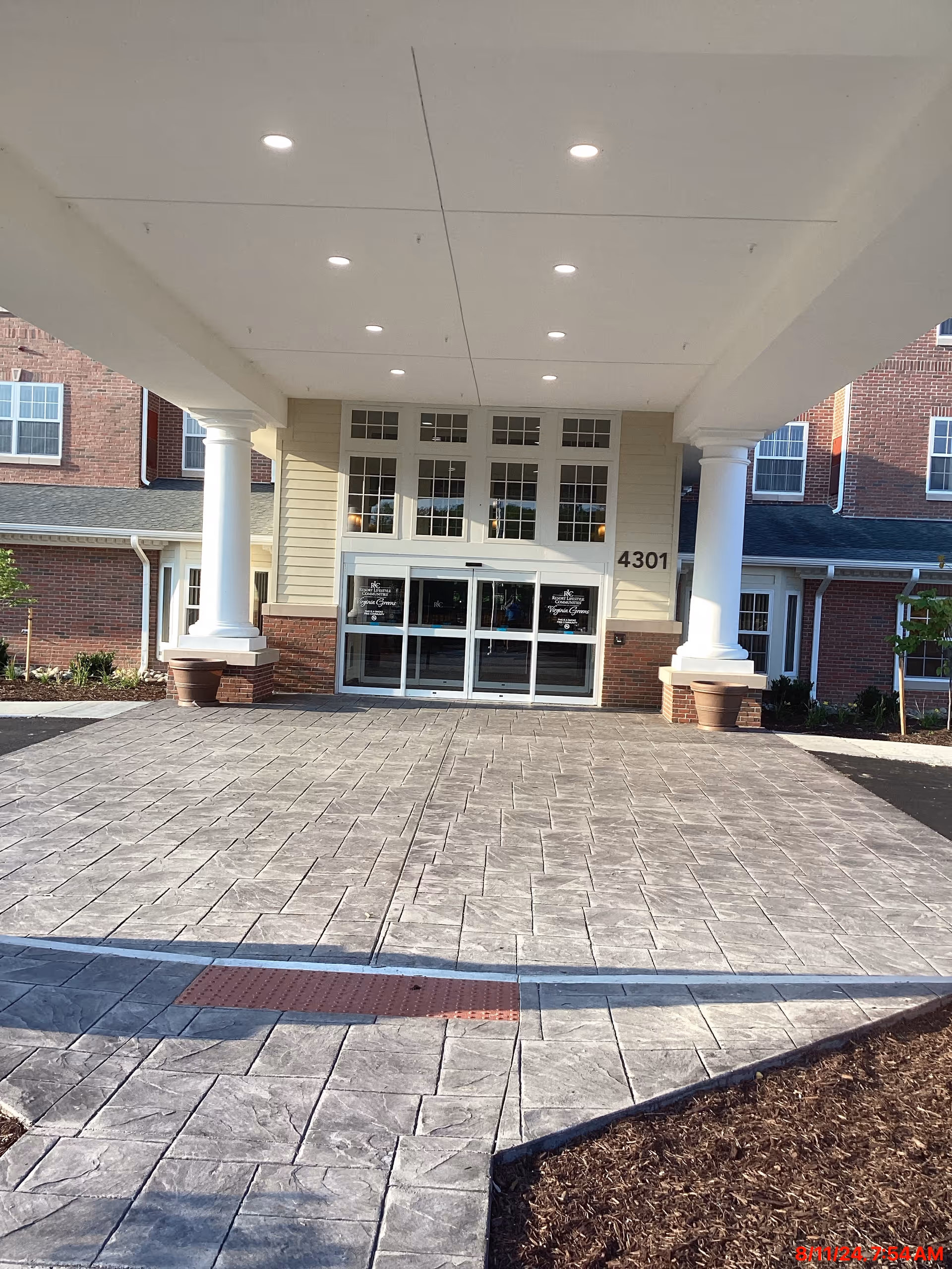 Entrance of Virginia Greens Retirement Community showing a covered driveway with large white columns, automatic glass doors, and brick and beige siding exterior walls. The building number 4301 is visible on the right side of the entrance.