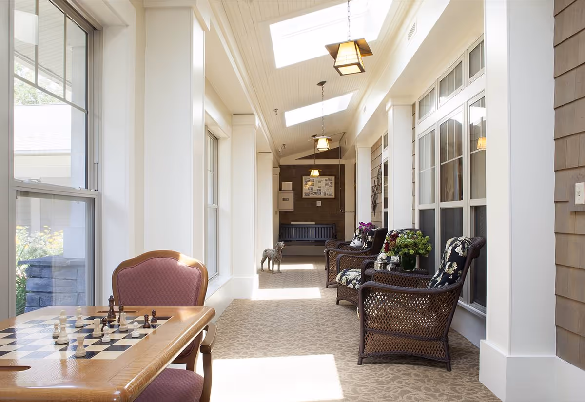 A bright indoor sunroom with large windows and skylights. The room features a chess table with pieces set up, a pink cushioned chair, and a seating area with wicker chairs that have floral cushions. There are potted plants on a small table between the chairs and a small dog standing in the background near a bench.