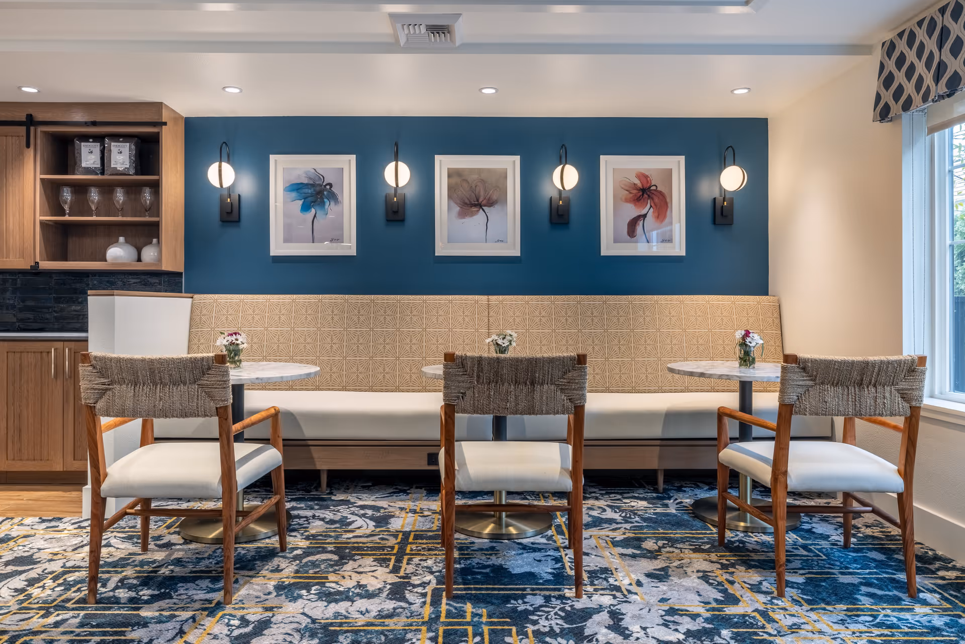 A cozy dining area in a senior living facility featuring three small round tables with beige cushioned chairs and a long beige upholstered bench against a blue accent wall. The wall is decorated with three framed floral paintings and four modern wall sconces with round white lights. To the left, there is a wooden cabinet with glassware and decorative items, and to the right, a window with patterned valance lets in natural light. The floor is covered with a patterned blue and yellow carpet.