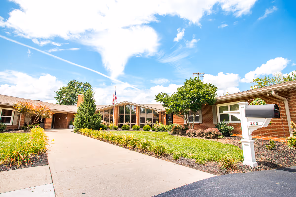 Exterior view of Fountain Circle Care & Rehabilitation Center showing a single-story brick building with large windows, a driveway leading to the entrance, a mailbox labeled 200, green shrubs, trees, and a bright blue sky with scattered clouds.