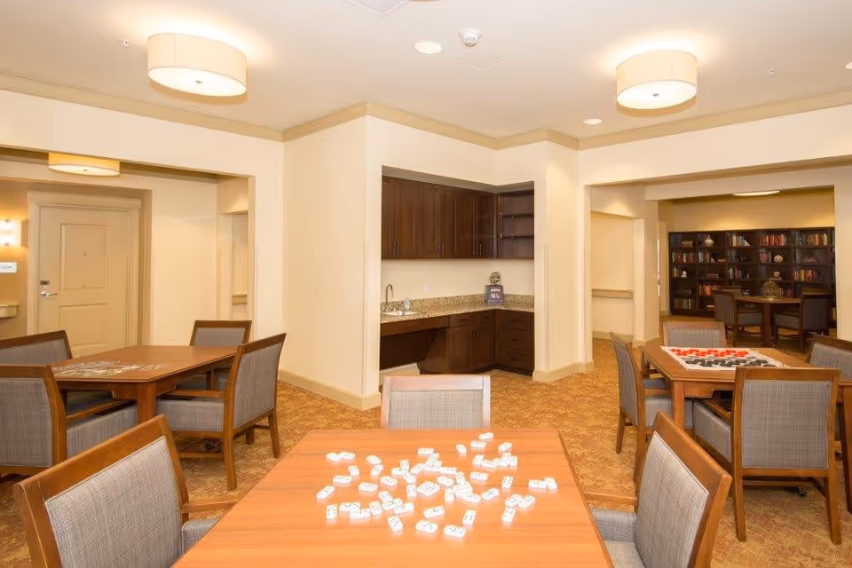 A well-lit common area in an assisted living facility featuring multiple wooden tables with chairs. One table has dominoes scattered on it, and another has a checkers game set up. In the background, there is a small kitchenette with dark wood cabinets and a granite countertop. Beyond the kitchenette, a room with bookshelves filled with books and decorative items is visible.