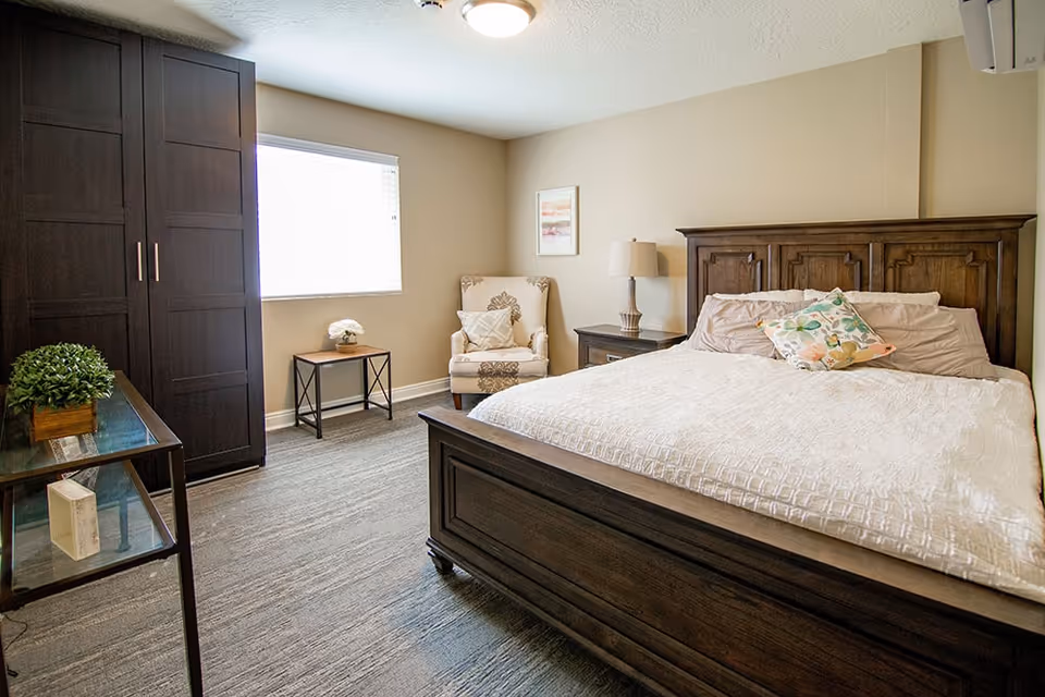 A bright bedroom with a large wooden bed featuring a white quilt and decorative pillows. There is a wooden nightstand with a lamp beside the bed, a patterned armchair in the corner, a small table with a flower vase near a window with blinds, and a dark wooden wardrobe. The room has beige walls and carpeted flooring.