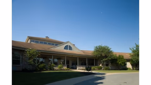 Single-story senior living building with a covered entrance, landscaped shrubs, and a clear blue sky.