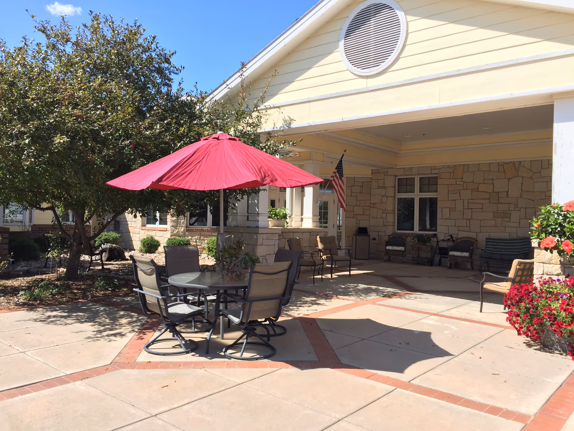 Outdoor patio area with a round glass table and four chairs under a large red umbrella. The patio is adjacent to a building with stone and yellow siding, featuring an American flag near the entrance. There are trees, shrubs, and flower pots with red and pink flowers around the patio.