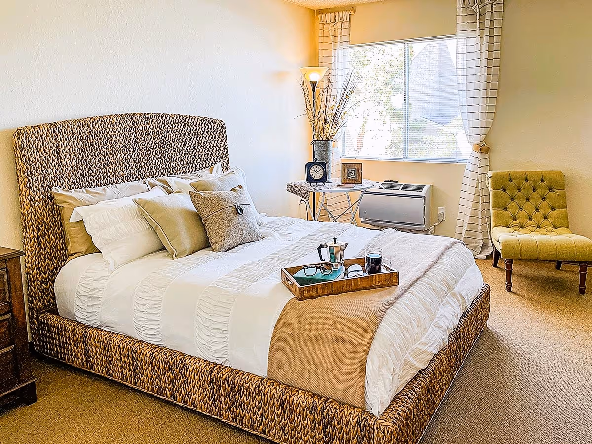 A cozy bedroom with a woven rattan bed frame and a neatly made bed with white and beige bedding. On the bed is a wooden tray holding a coffee pot, a glass of coffee, a pair of glasses, and a book. Next to the bed is a small wooden nightstand. A window with white curtains lets in natural light, and a green upholstered chair is placed near the window. A floor lamp and a small table with decorative items are also visible in the corner.