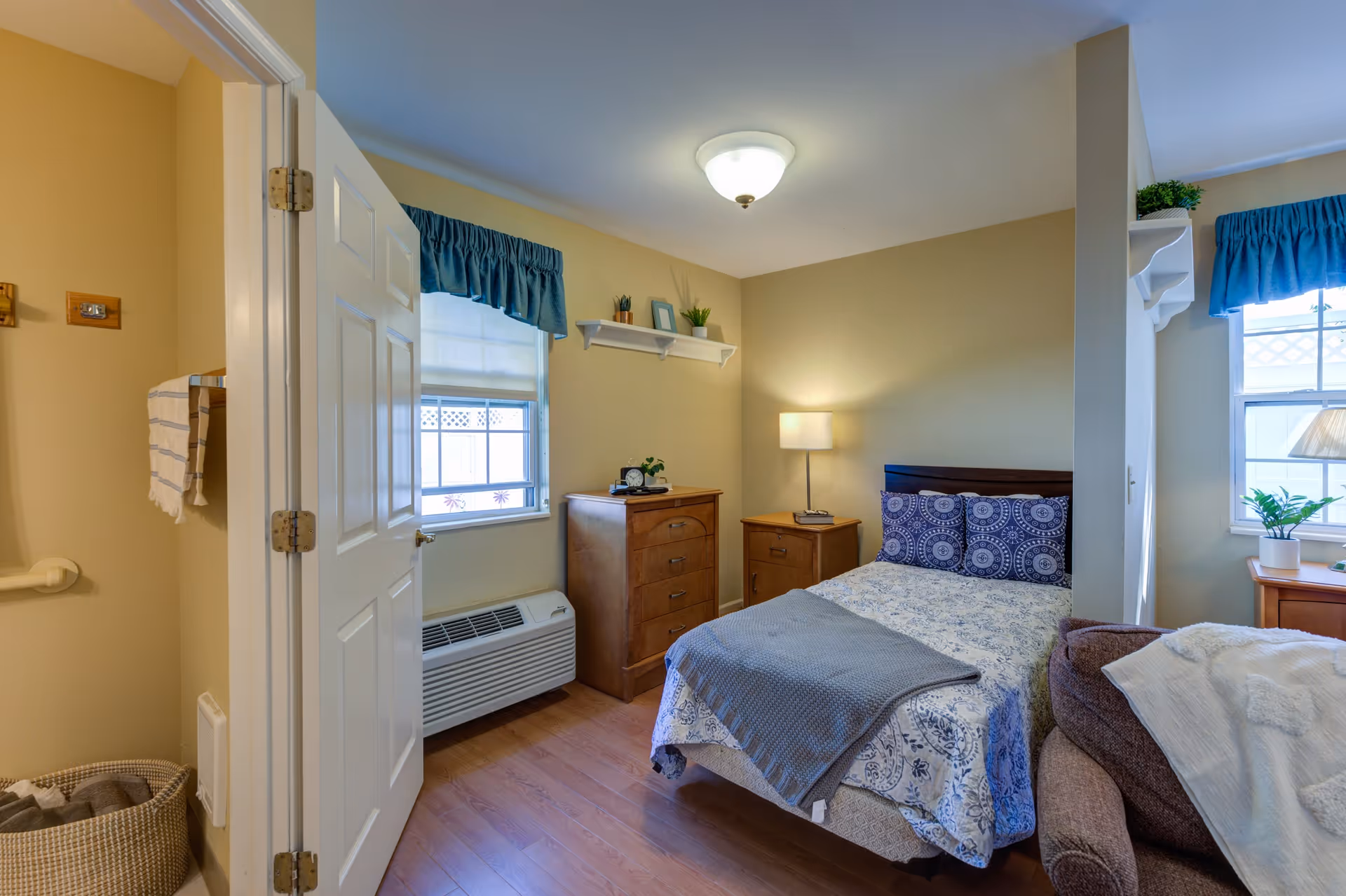 A cozy bedroom in a senior living facility with a single bed covered in patterned bedding and a gray throw blanket. The room has wooden flooring, a wooden dresser and nightstand with a lamp, and two windows with blue valances. There is a brown armchair with a white blanket draped over it, and an open door leading to a bathroom with a towel hanging on a rack and a laundry basket on the floor.