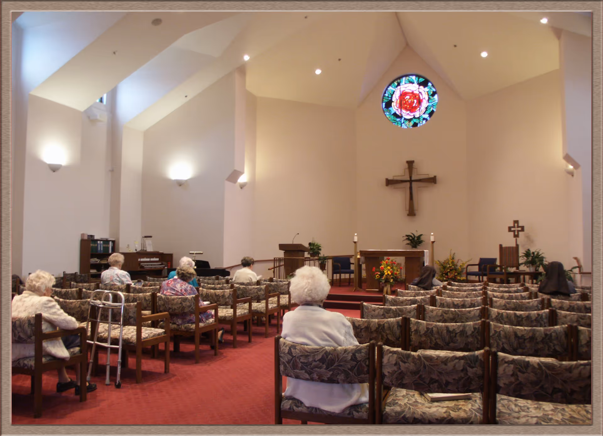 Interior of a chapel with elderly people seated on floral-patterned chairs facing an altar with a wooden cross on the wall and a stained glass window above it. The room has high ceilings with soft lighting and red carpeted floor.