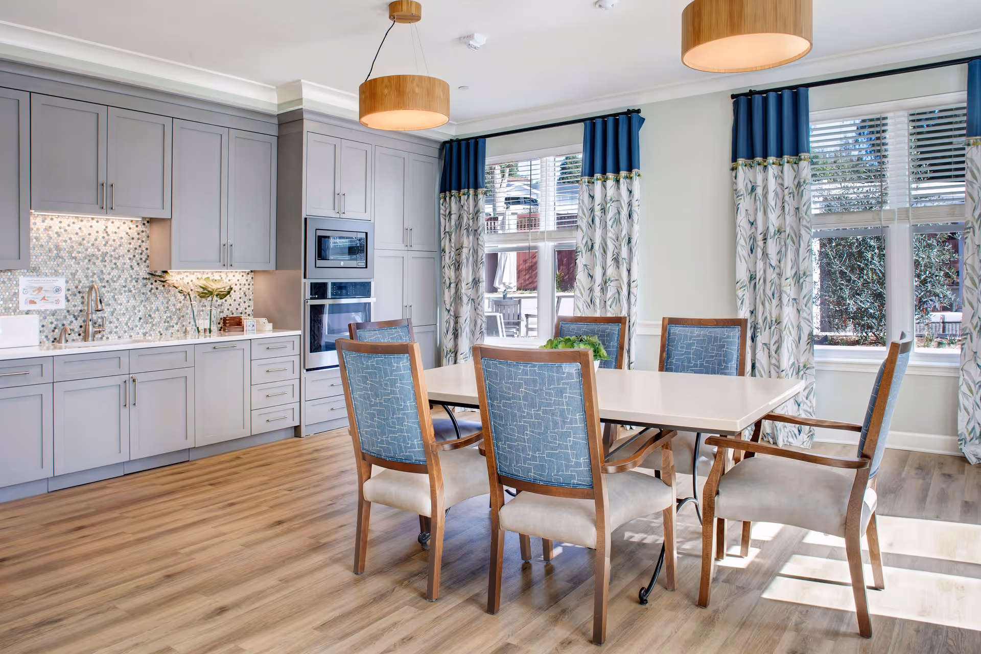 A bright dining area with a rectangular table surrounded by six wooden chairs with blue patterned backs and beige seats. The room features light wood flooring, gray cabinetry with built-in microwave and oven, a mosaic tile backsplash, and large windows with blue and white patterned curtains letting in natural light. Two round pendant lights hang from the ceiling.