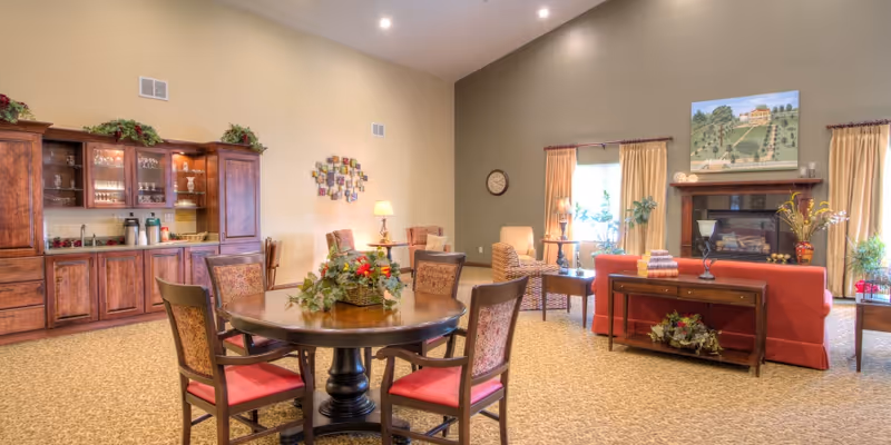 A spacious and warmly decorated living room area with a round wooden table and four chairs with red cushions in the foreground. Behind the table is a wooden cabinet with glassware and coffee supplies. The room features a fireplace with a painting above it, two large windows with beige curtains, and several comfortable armchairs and sofas arranged around the fireplace. The walls are painted in soft yellow and green tones, and the floor is carpeted.