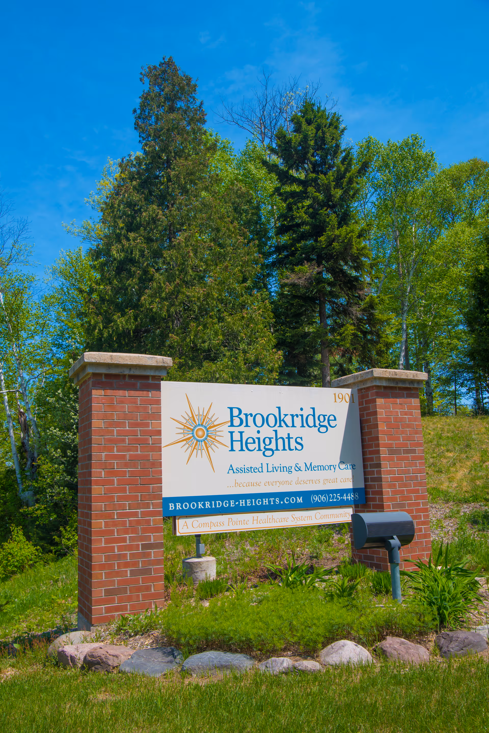 Entrance sign for Brookridge Heights assisted living set between two brick pillars with trees and blue sky behind.