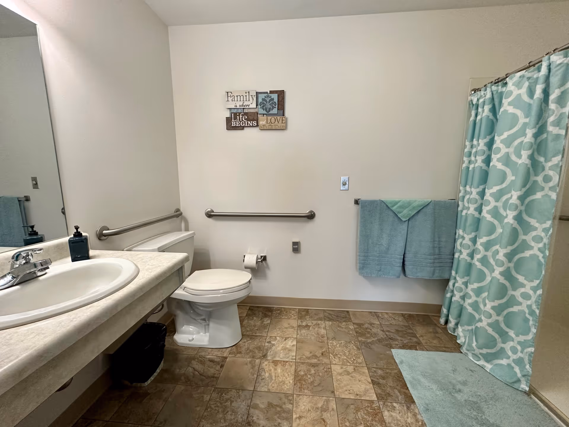 A clean bathroom with a white toilet, a sink with a countertop, a large mirror, and a shower with a blue and white patterned curtain. There are two blue towels hanging on a towel rack and a blue bath mat on the floor. The walls are light-colored with a decorative sign that reads 'Family is where Life Begins and Love Never Ends.' The floor has brown and beige tiles.