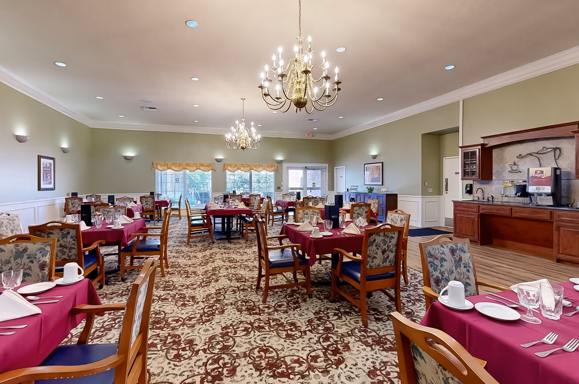 A spacious dining room with multiple tables covered in burgundy tablecloths, each set with plates, glasses, napkins, and silverware. The chairs have wooden frames with floral upholstery. The room features patterned carpet, light green walls with white wainscoting, and two chandeliers hanging from the ceiling. Large windows with valances allow natural light to enter, and there is a counter area with a coffee machine and sink on the right side.