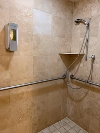 Shower area with beige tiled walls and floor, equipped with a handheld showerhead, grab bars on two walls, and a wall-mounted soap dispenser.