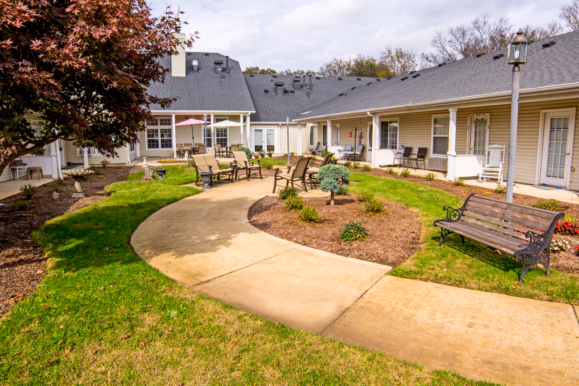 Sunny courtyard with a curved concrete walkway, patio chairs, benches and landscaping in front of a one-story residential building.