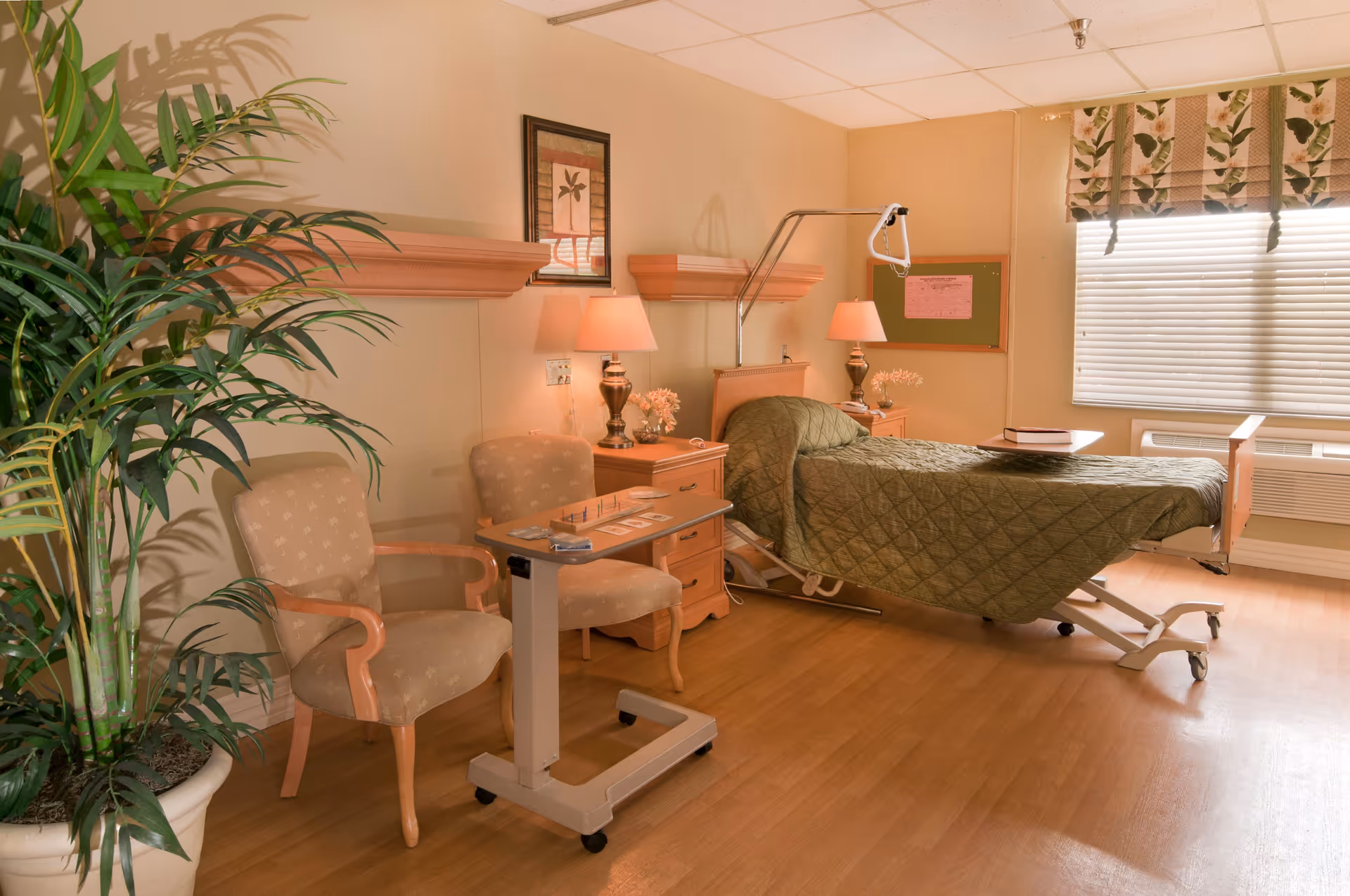 A well-lit patient room in a healthcare facility with a hospital bed covered in a green quilt, two beige armchairs, a small overbed table with a game on it, two bedside tables with lamps and flowers, a large potted plant, a window with blinds and floral valance, and a bulletin board on the wall.