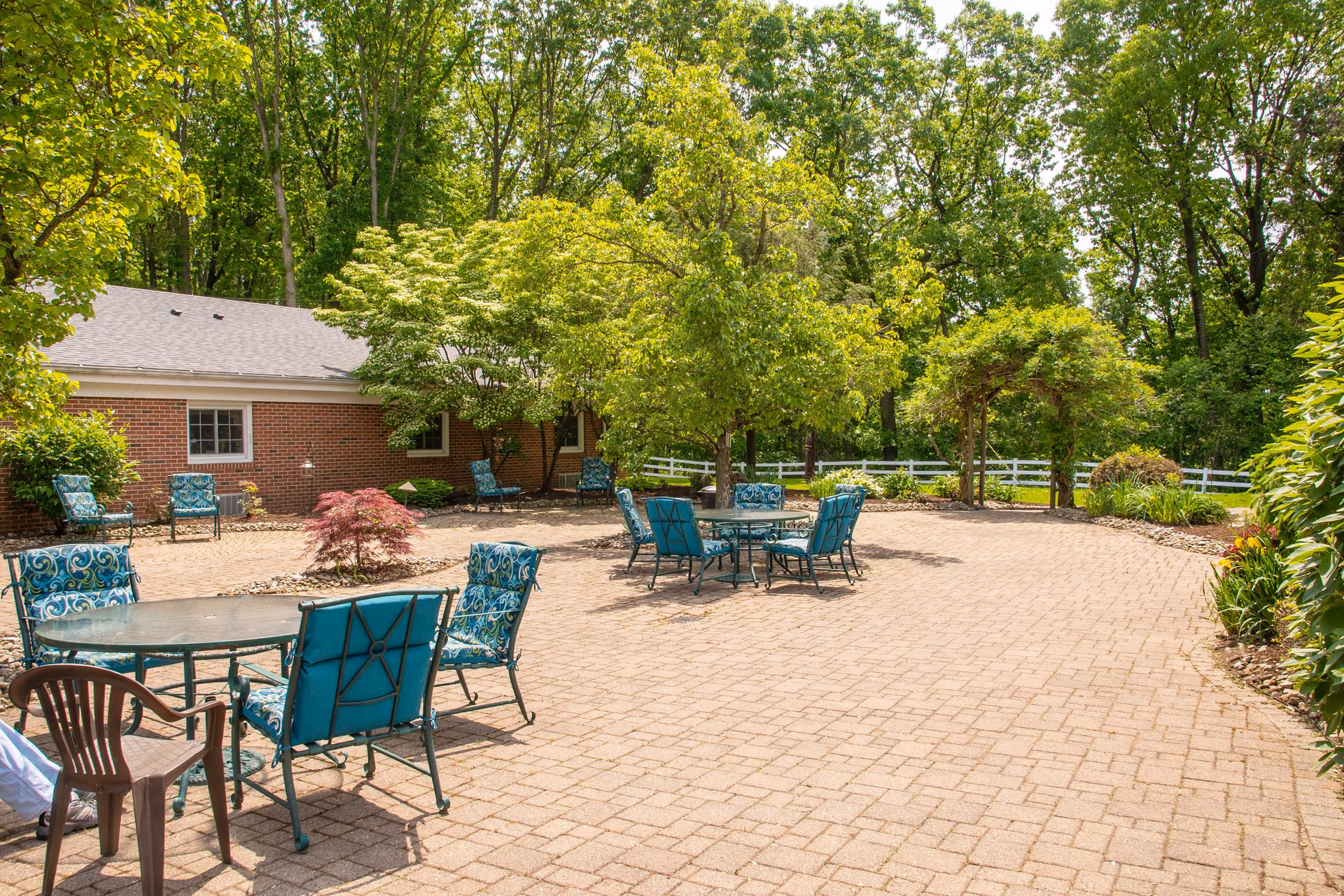 Outdoor patio area with several metal tables and chairs with blue cushions, surrounded by trees and greenery. A brick building is visible in the background along with a white fence and an arbor covered in plants.