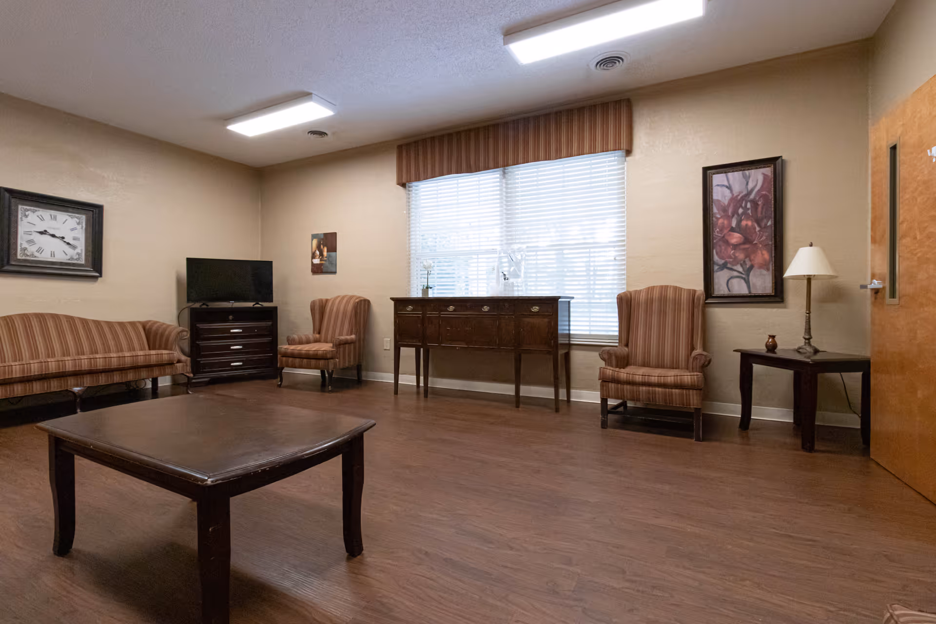 A cozy living room with wooden flooring, featuring a striped sofa and two matching armchairs. There is a dark wooden coffee table in the center, a TV on a small cabinet, a large window with blinds, a sideboard beneath the window, a framed floral painting, a wall clock, and a side table with a lamp next to a wooden door.