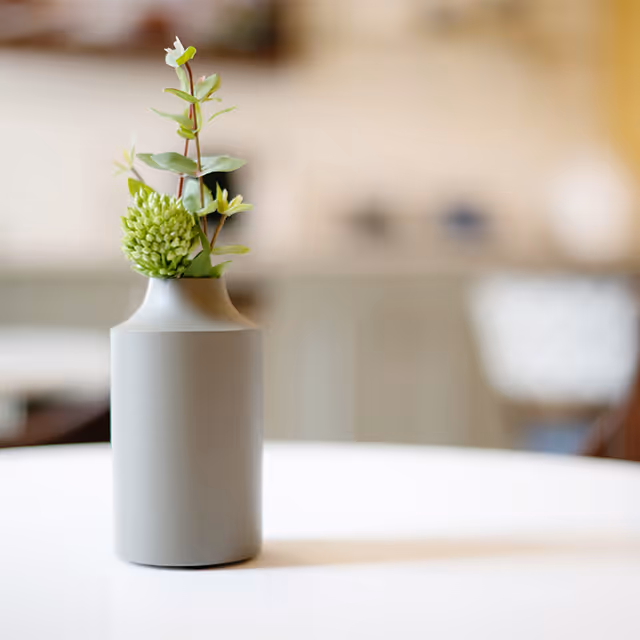 A small gray vase with green foliage and a green flower bud placed on a white table with a blurred background of an indoor setting.