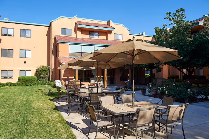 Outdoor patio area at Sandia Vista Senior Living with multiple tables and chairs under large beige umbrellas, surrounded by green grass, trees, and a multi-story building in the background under a clear blue sky.