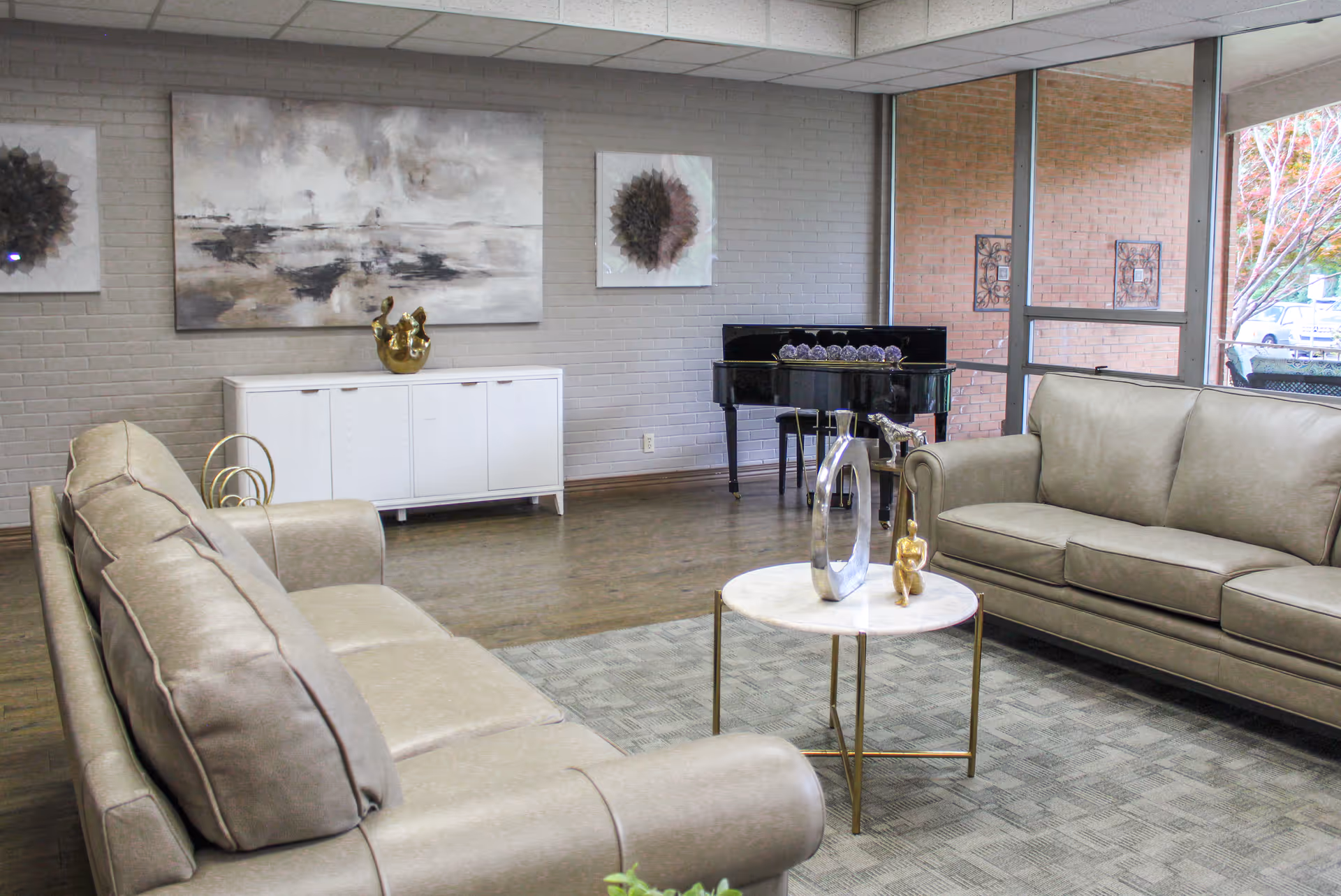 A modern living room with two beige leather sofas facing each other, a small round coffee table with decorative sculptures, a white cabinet against a gray brick wall, abstract artwork hanging above the cabinet, and a black grand piano near large windows showing an outdoor view.