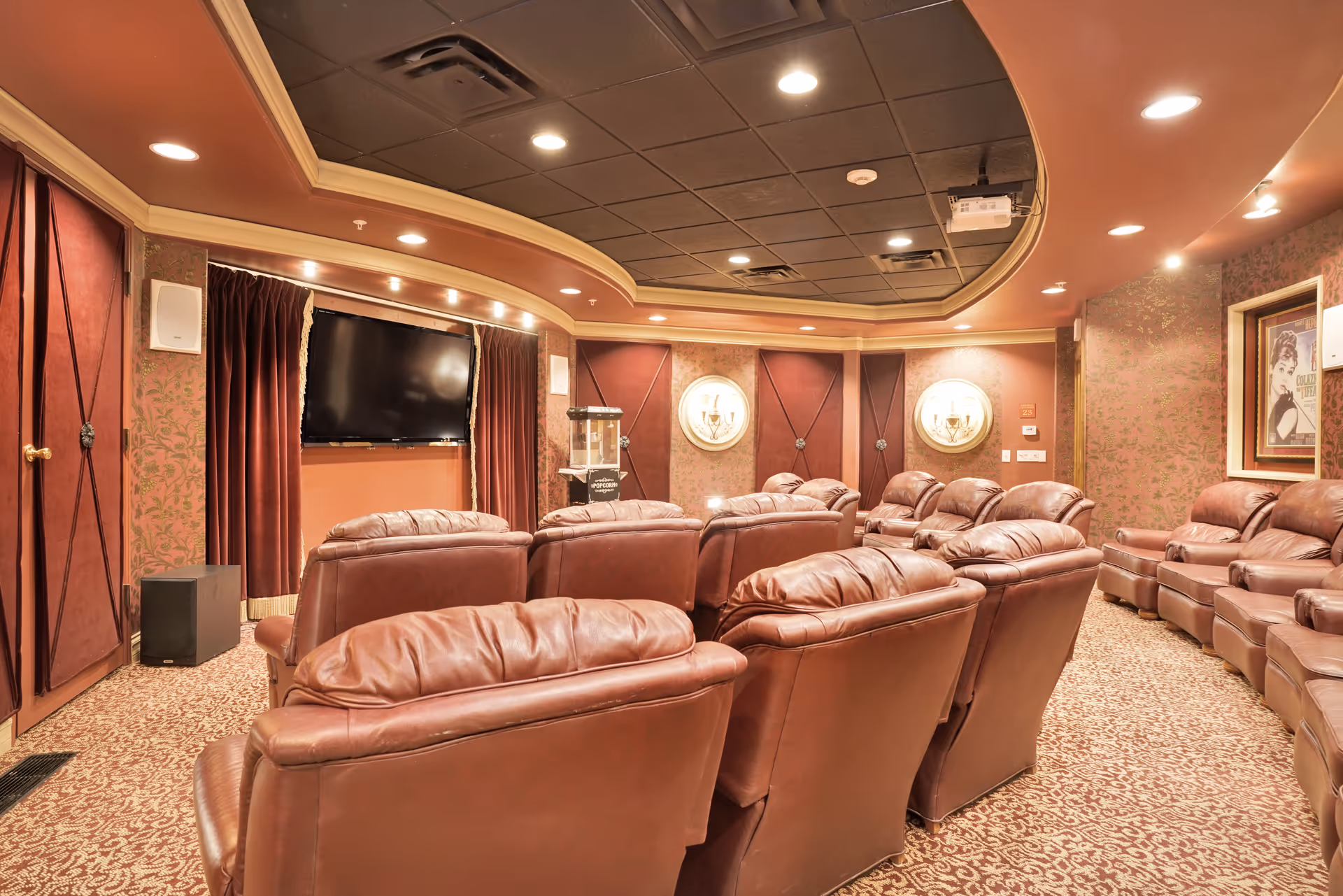 A cozy home theater room with multiple rows of brown leather recliner chairs facing a large flat-screen TV mounted on the wall. The room features warm lighting, patterned carpet, decorative wall sconces, and framed artwork on the walls.