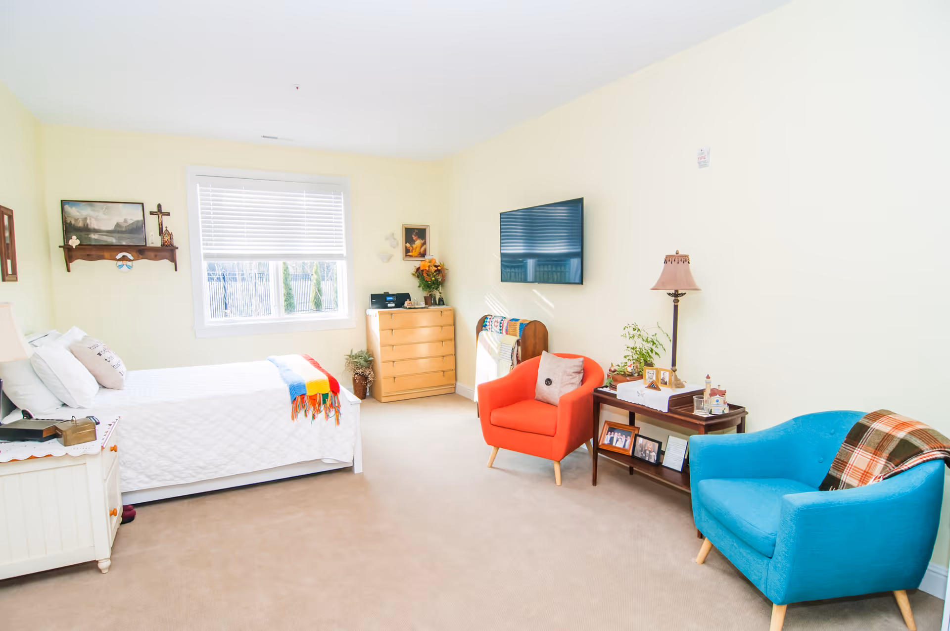 A bright and cozy senior living bedroom with a white bed covered in a quilt and colorful blanket, two armchairs in red and blue, a wooden dresser, a wall-mounted TV, and a side table with a lamp and framed photos. The room has light yellow walls and a window with white blinds letting in natural light.