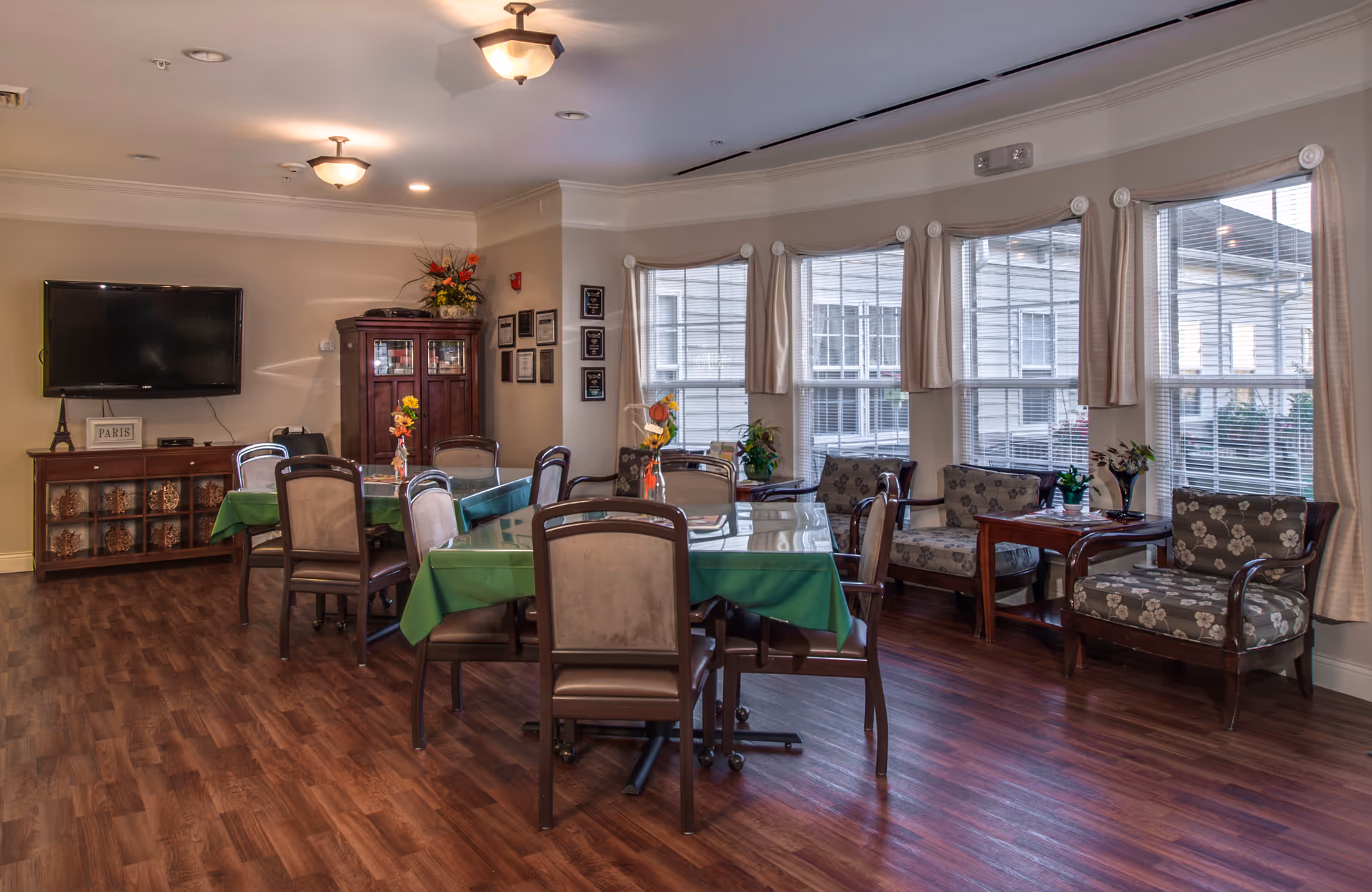 A senior living facility common area with multiple tables covered in green tablecloths and surrounded by chairs. There is a wooden cabinet with glass doors and a flat-screen TV mounted on the wall. Large windows with beige curtains let in natural light, and there are several cushioned armchairs with floral patterns arranged near the windows. The room has wood flooring and ceiling lights.