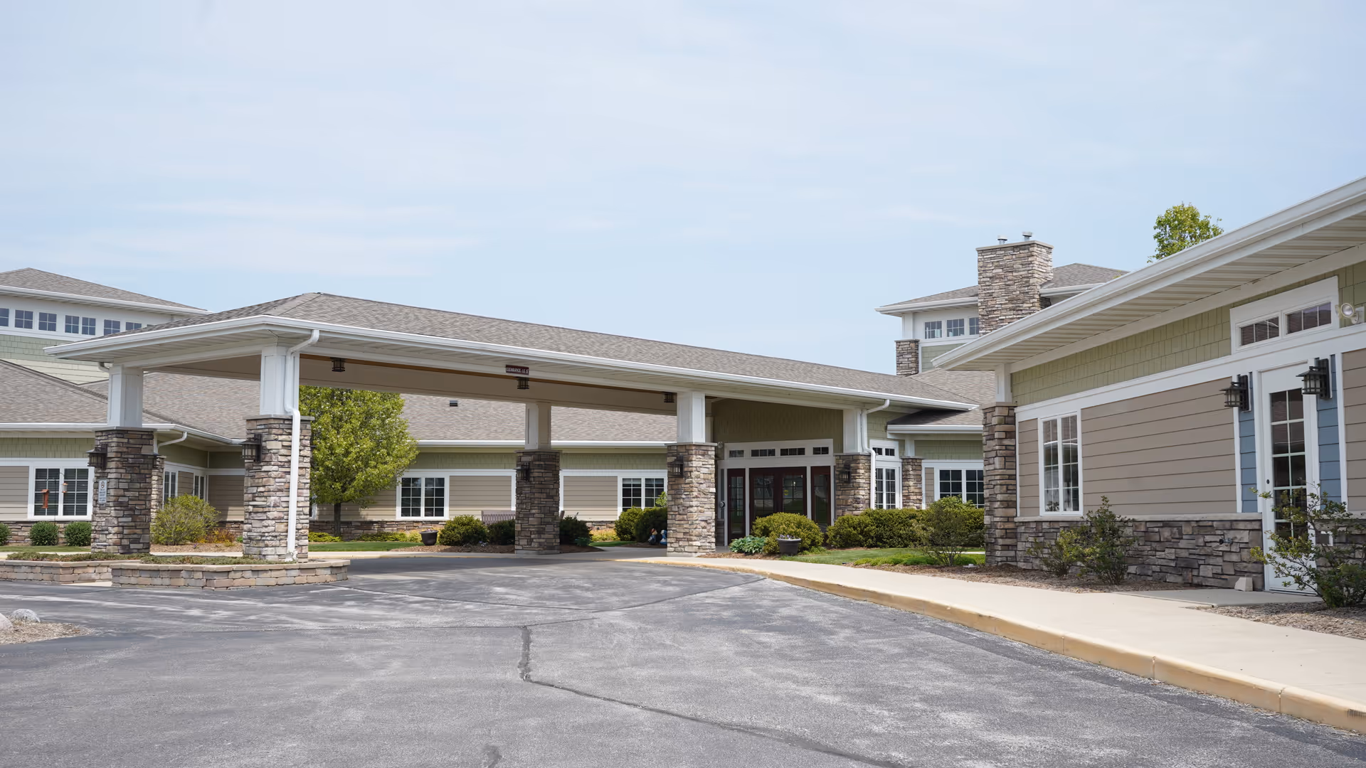 Exterior view of Pine Haven Christian Communities Oostburg Campus showing a covered entrance supported by stone pillars, with beige siding and stone accents on the building, surrounded by landscaping and a paved driveway.