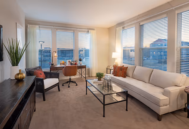 A bright and cozy living room with large windows covered by white blinds, a beige sofa with decorative pillows, a glass coffee table with a small plant and decorative items, a wooden chair with a cushion, a desk with a brown office chair, and a lamp on a side table. The room has beige carpet and neutral-colored walls.