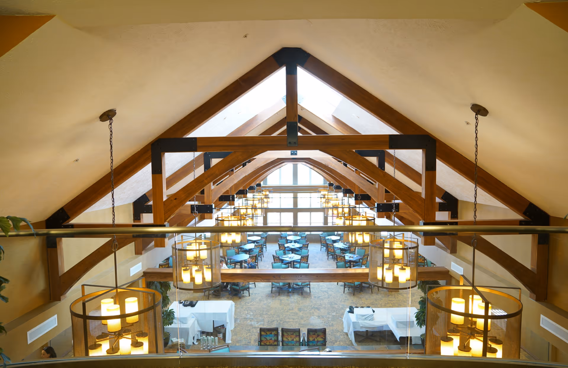 Interior view of a large dining room with high vaulted ceilings supported by wooden beams. The room is filled with multiple round tables and chairs arranged neatly. Several hanging light fixtures with multiple bulbs provide warm lighting. The space has large windows at the far end allowing natural light to enter.