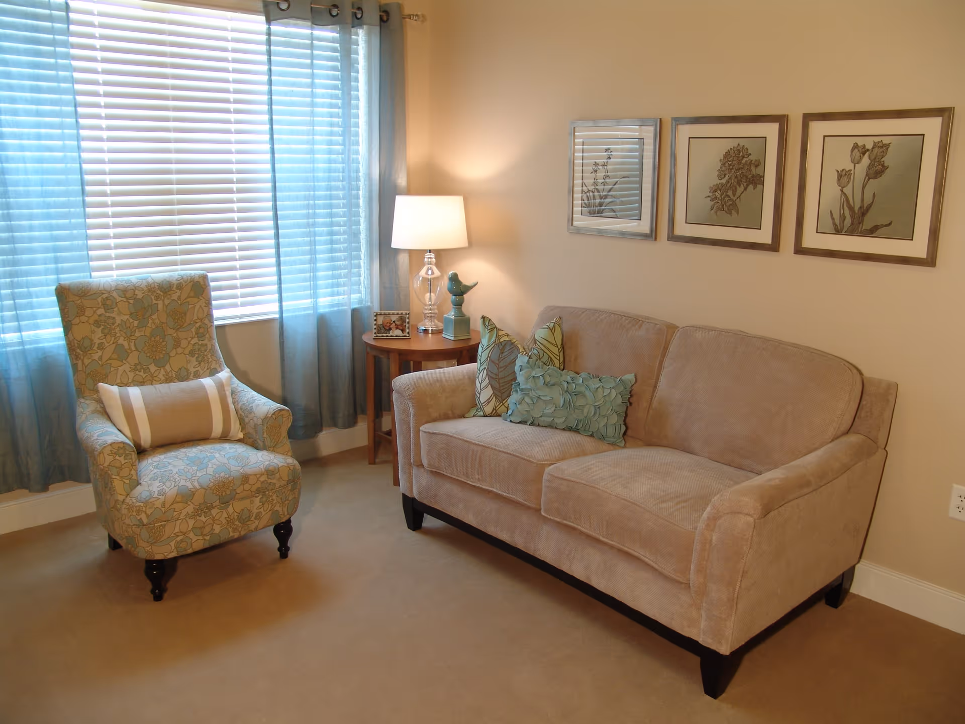 A cozy living room with a beige sofa adorned with three decorative pillows, a floral patterned armchair with a striped lumbar pillow, a wooden side table with a lamp, a small bird figurine, and a framed photo. The room has beige walls with three framed botanical prints and a window with blue curtains and white blinds.