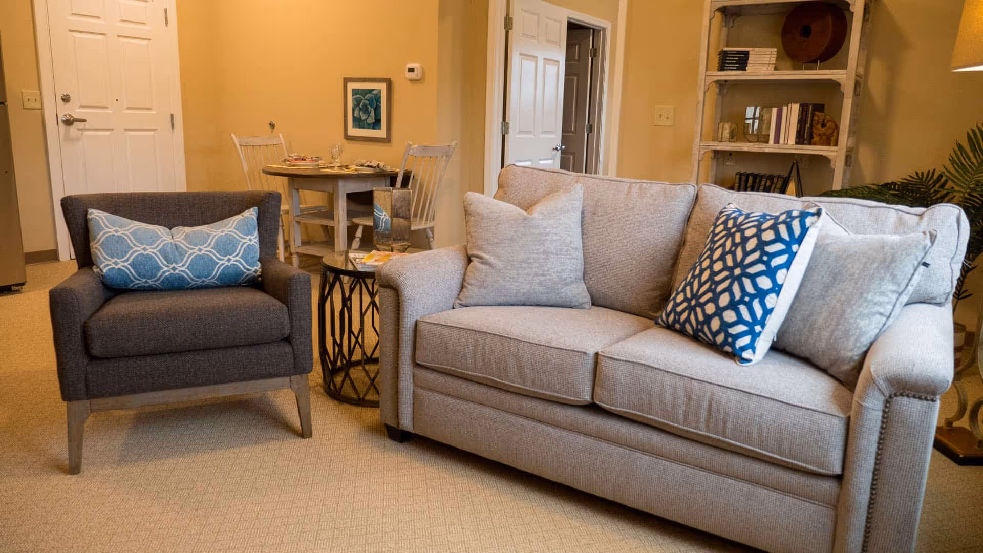 A cozy living room area featuring a beige sofa with three cushions, including one with a blue and white geometric pattern, and a dark gray armchair with a blue patterned pillow. Behind the seating area is a small round dining table with two chairs, set with plates and glasses. A white door and a bookshelf with books and decorative items are visible in the background.