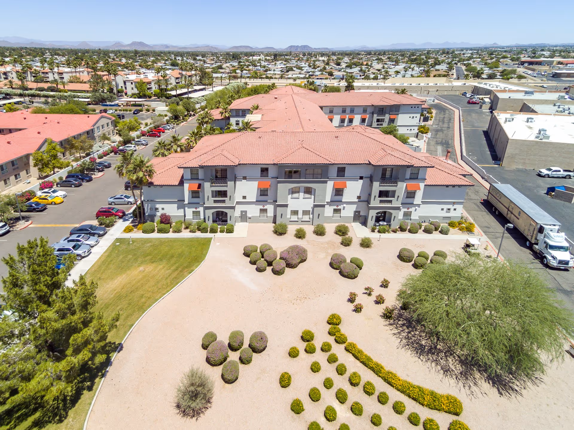 Aerial view of a senior living facility with a large building featuring red-tiled roofs and white walls. The foreground shows landscaped grounds with bushes, small trees, and a sandy area. There are parking lots with cars on the left side and a truck parked on the right side. The background includes more buildings and a distant mountain range under a clear sky.