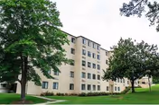 Beige multi-story residential building with windows set in a grassy, tree-lined lawn under a cloudy sky.