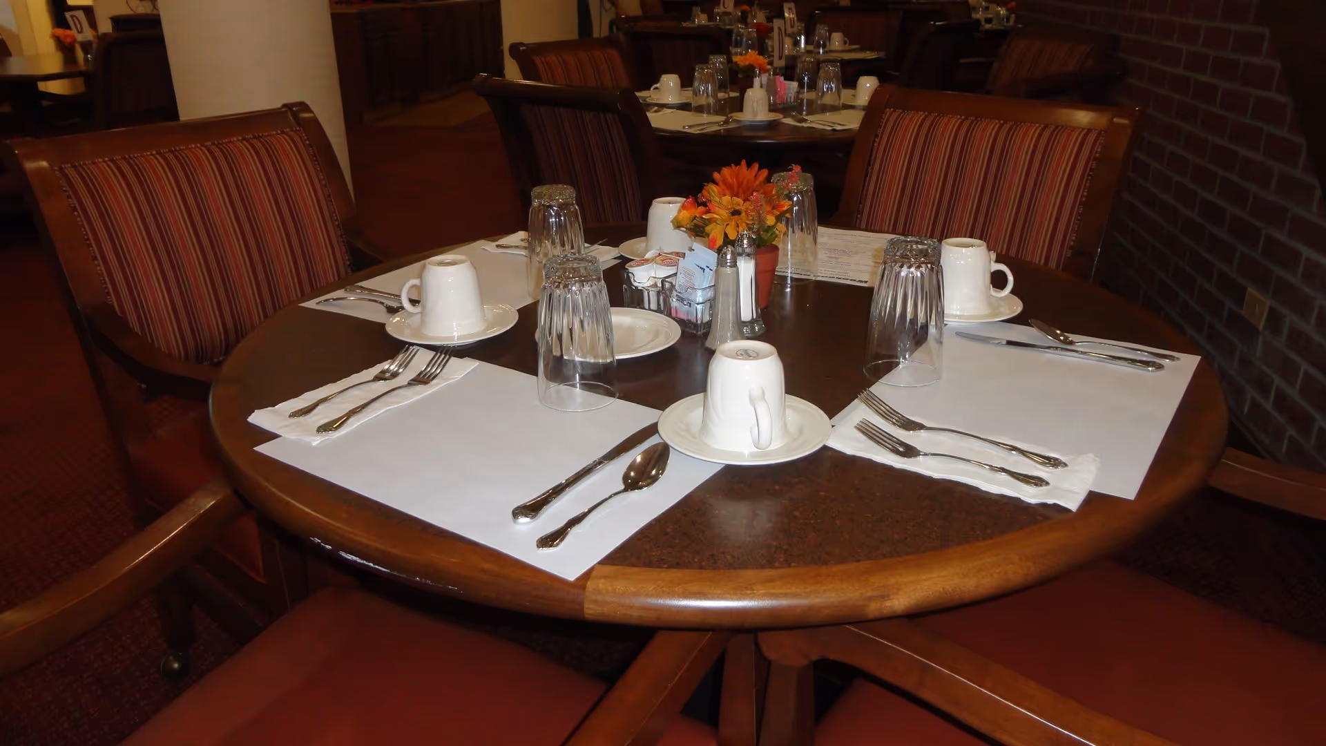 Round dining table set for four with cups, glasses, silverware, placemats and a small flower centerpiece in a dining room.