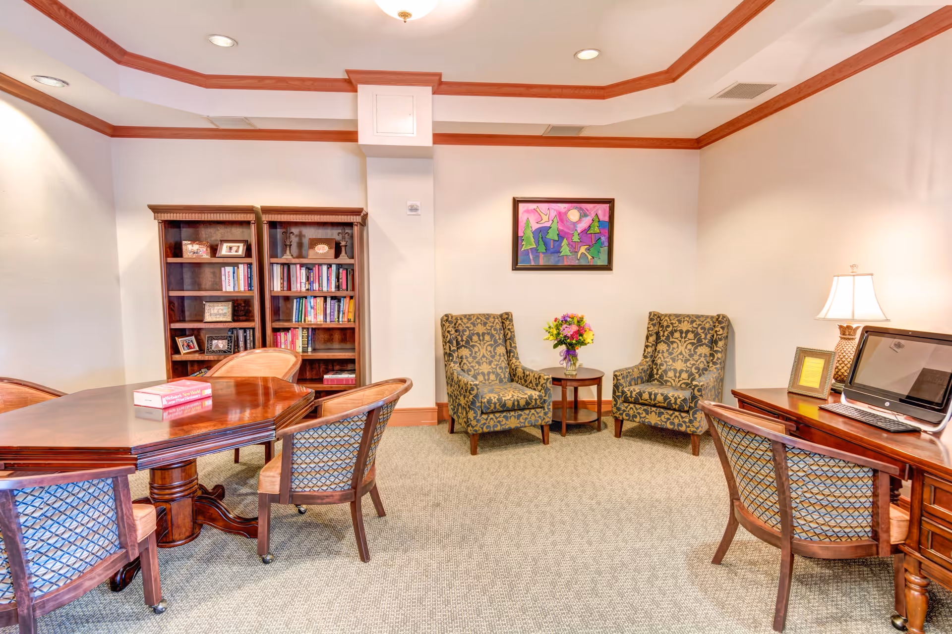 A cozy interior room with a wooden table surrounded by chairs, two patterned armchairs with a small round table holding a vase of flowers between them, a wooden desk with a computer and lamp, and two wooden bookshelves filled with books and framed photos. The walls are light-colored with crown molding and a colorful painting hangs above the armchairs.