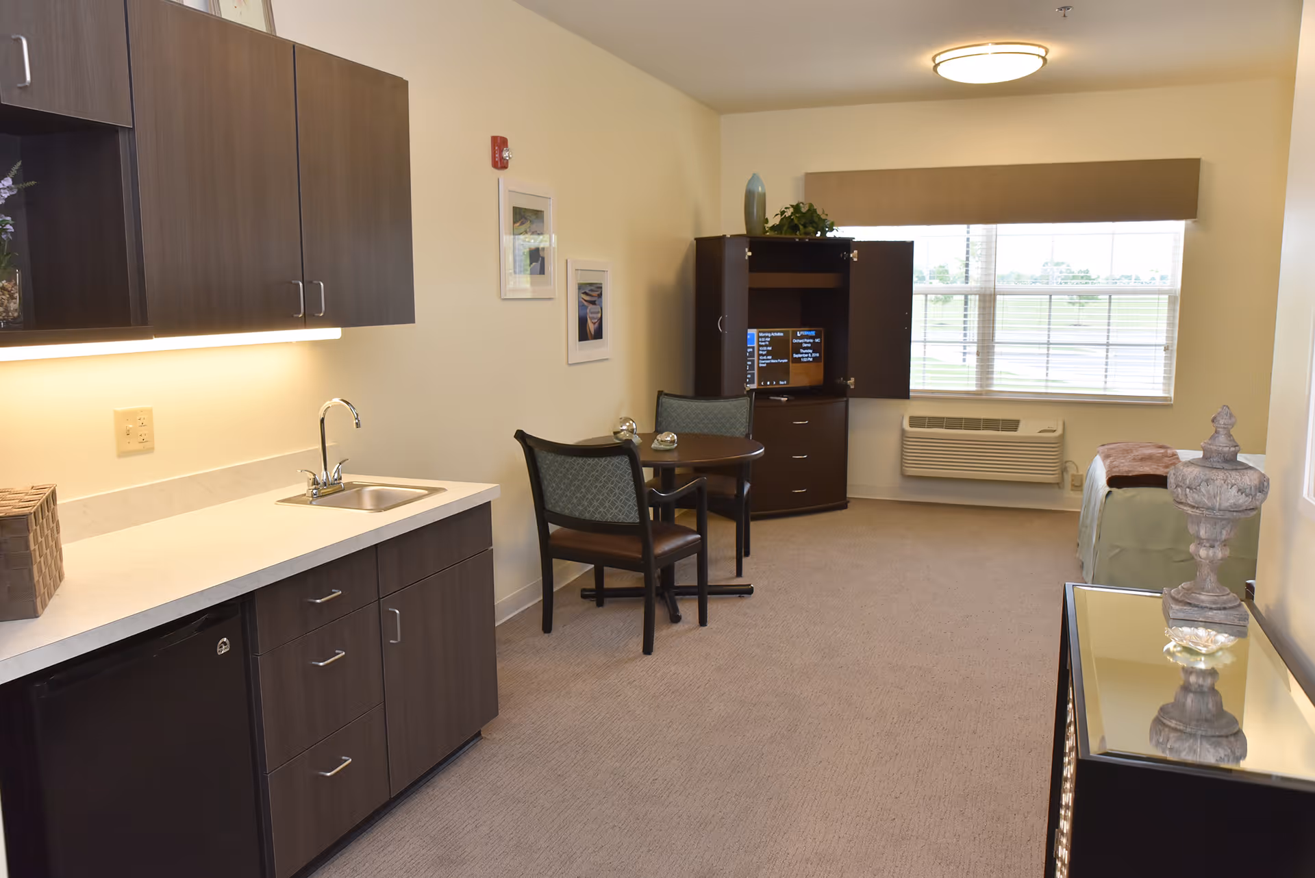 Bright studio-style senior living interior with a kitchenette on the left, a small dining table and TV cabinet in the center, and a bed near the window.