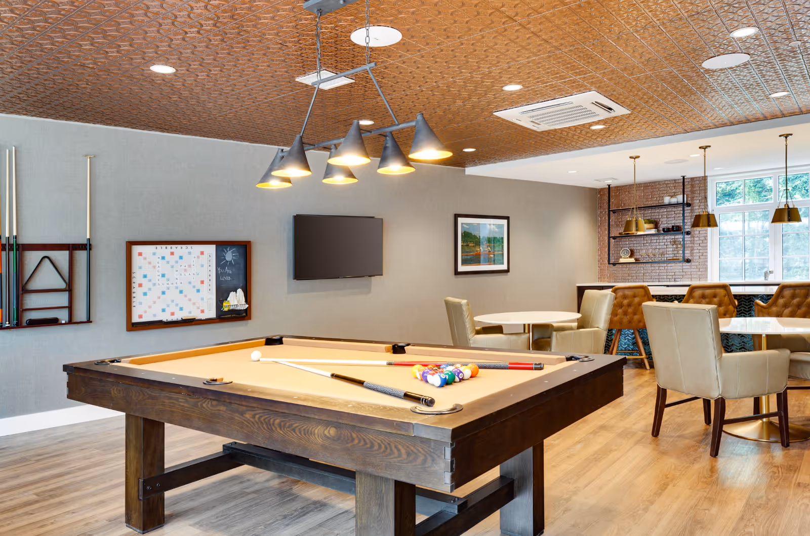 A bright common room with a pool table, seating and pendant lights in a senior living facility.