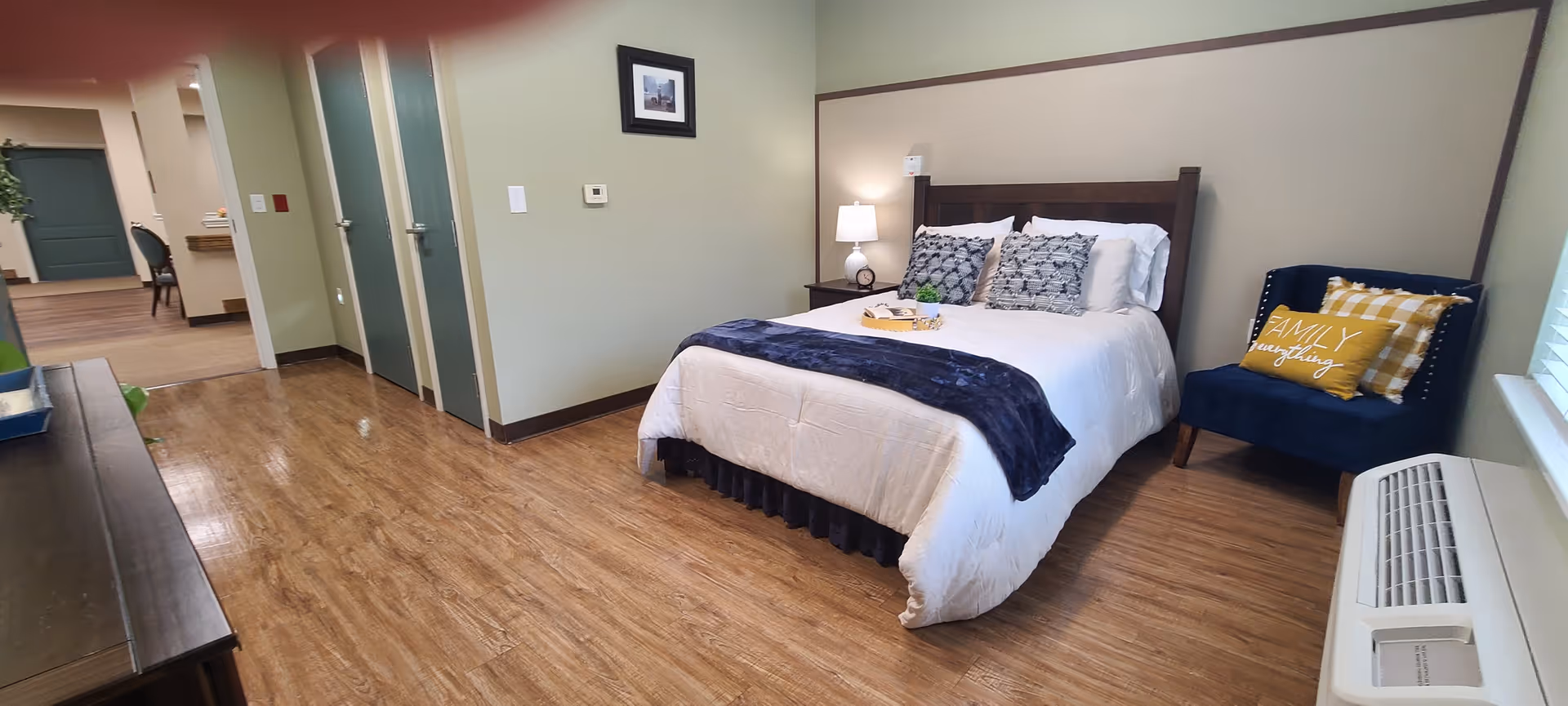 A neatly made bed with white bedding and decorative pillows in a room with wooden flooring. Next to the bed is a small nightstand with a lamp. A blue armchair with a yellow pillow that says 'FAMILY everything' is placed near a window with blinds. The room has light green walls and a thermostat on one wall. In the background, there is a hallway with doors and a glimpse of another room with chairs and a table.