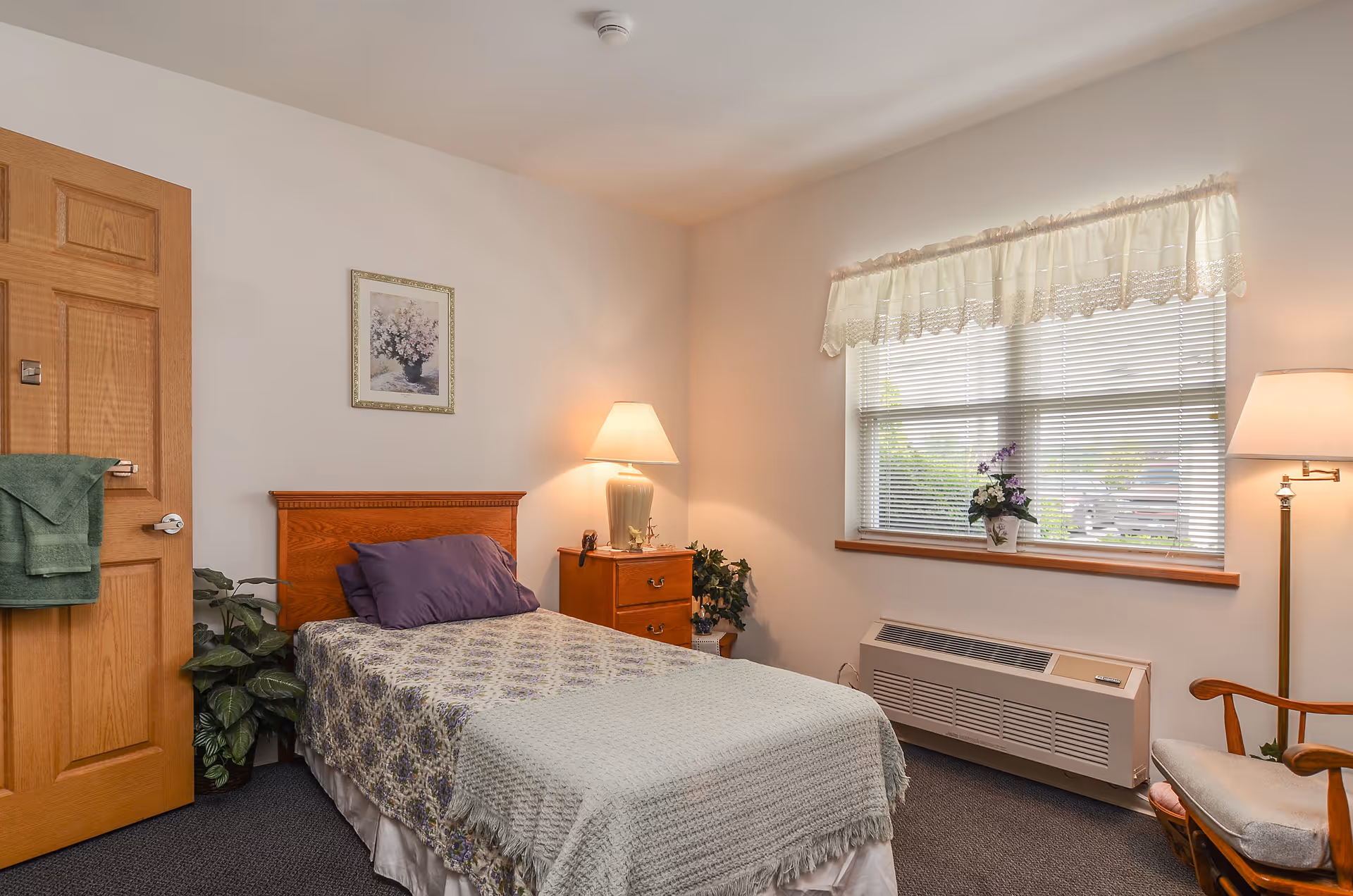 A cozy bedroom with a single bed covered in a patterned bedspread and a purple pillow. There is a wooden nightstand with a lamp and a small plant next to the bed. A wooden door with a green towel hanging on the handle is on the left side. A window with blinds and a lace valance lets in natural light, with a potted plant on the windowsill. A cushioned wooden chair and a floor lamp are positioned near the window.