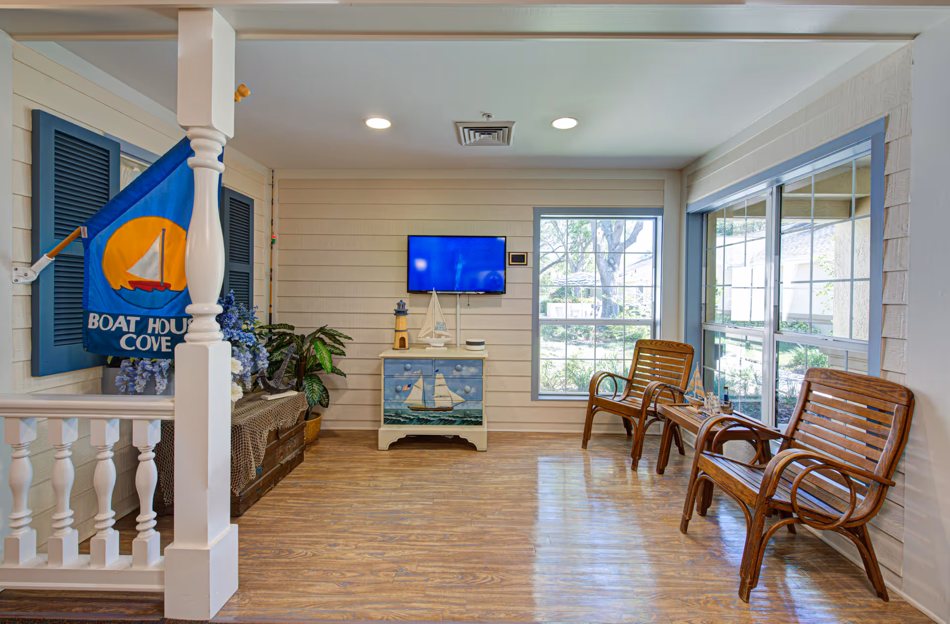 A cozy interior room with wooden flooring and light-colored walls. There are two wooden chairs with armrests and a small wooden table between them, decorated with small model ships. A flat-screen TV is mounted on the wall above a small cabinet painted with a sailboat design. A blue flag with a sailboat and the words 'Boat House Cove' hangs on the left side near a white railing and a decorative chest with plants and flowers.