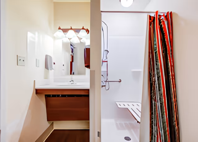 A bathroom area with a sink and mirror on the left side and a shower with a foldable bench and a striped shower curtain on the right side.
