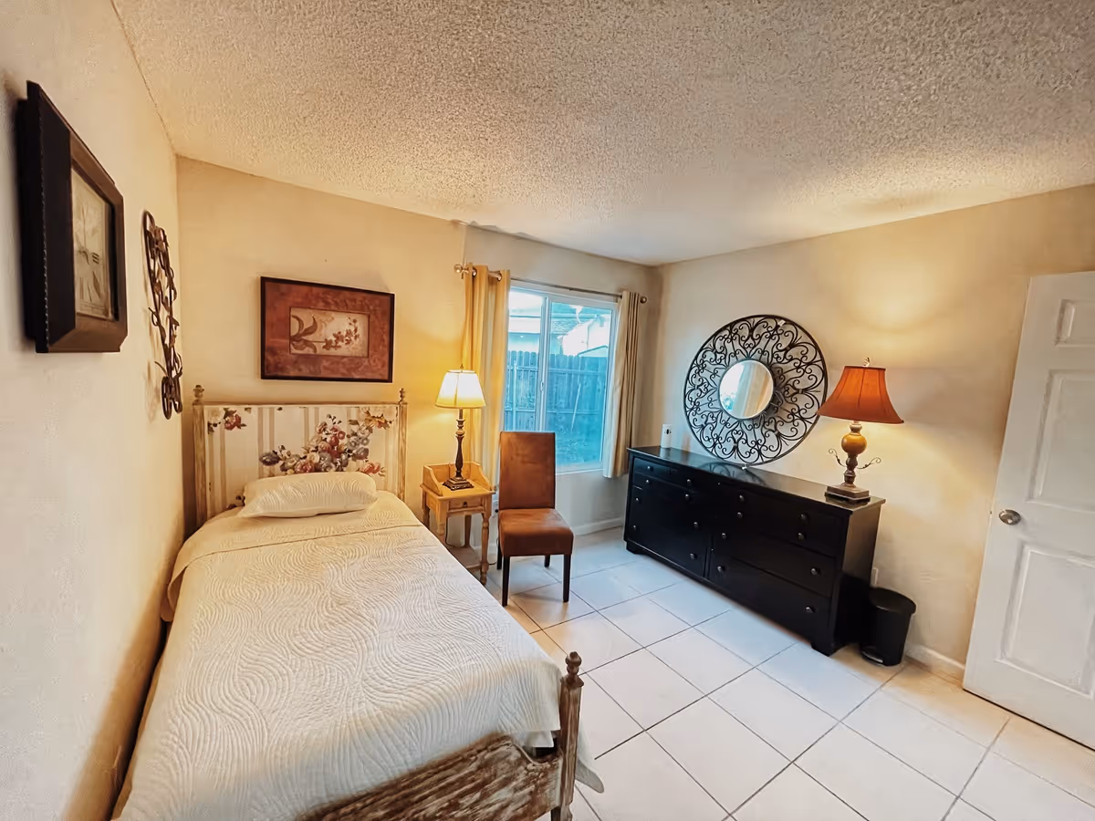 Small bedroom with a single bed, nightstand and lamp, a chair by a window, and a dresser topped by a decorative round mirror.