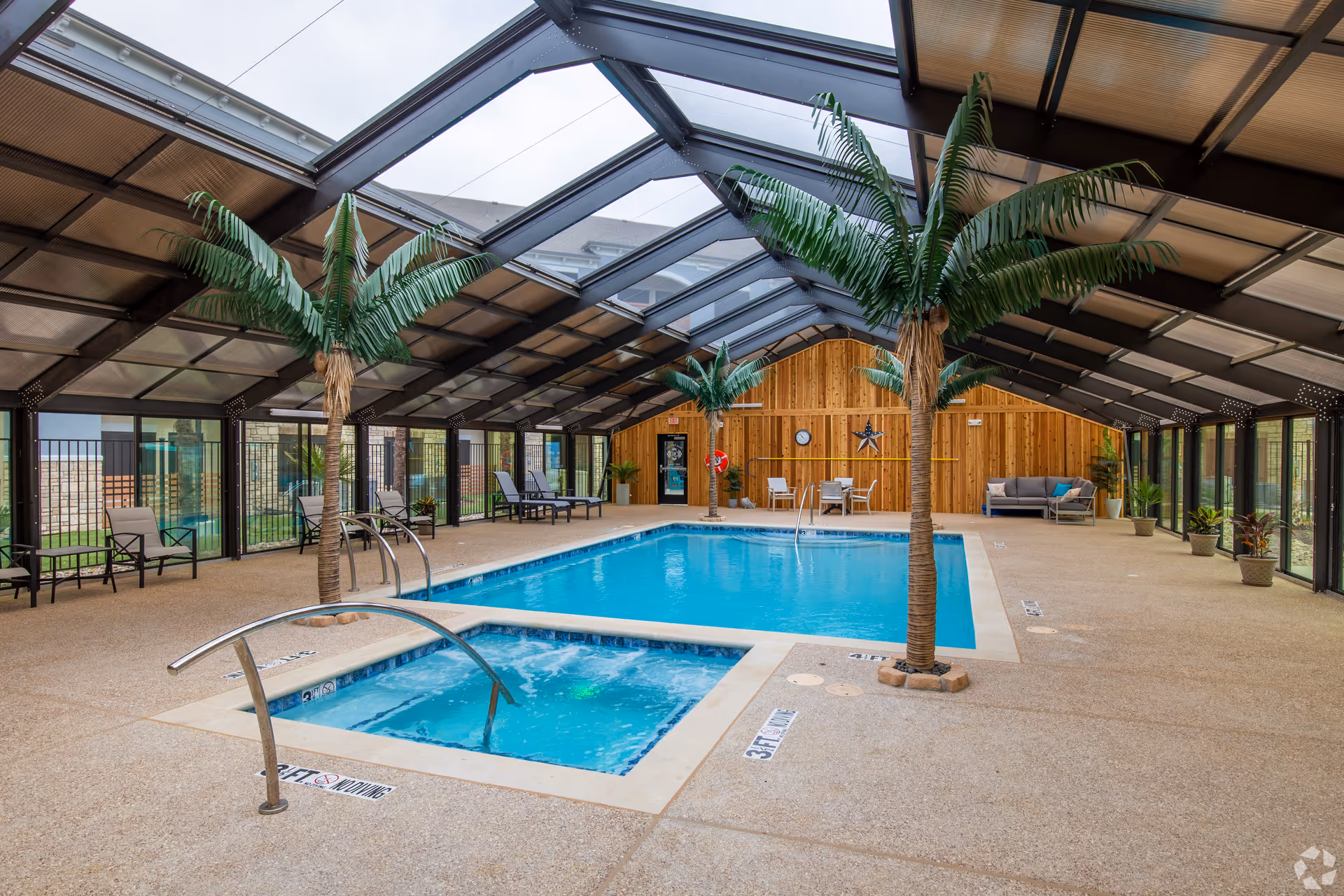 Indoor pool area with a hot tub in the foreground and a swimming pool behind it, surrounded by artificial palm trees, lounge chairs, potted plants, and a wooden wall with a clock and star decoration. The area is enclosed with glass walls and a transparent roof.