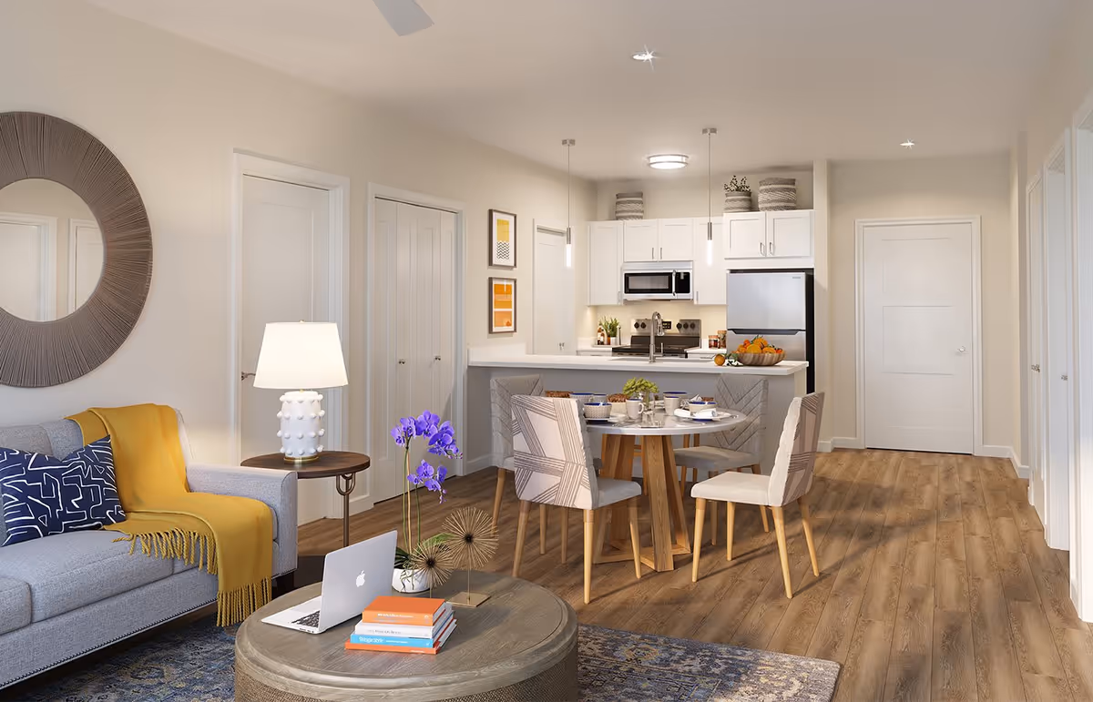 A modern open-concept living and dining area with a gray sofa adorned with a yellow throw and a blue patterned pillow. A round wooden coffee table holds a laptop, books, and a small plant with purple flowers. The dining area features a round table set for four with patterned chairs. The kitchen in the background has white cabinets, a stainless steel refrigerator, microwave, and stove, with pendant lights hanging above the counter. The room has light wood flooring and neutral-colored walls with minimal decor.