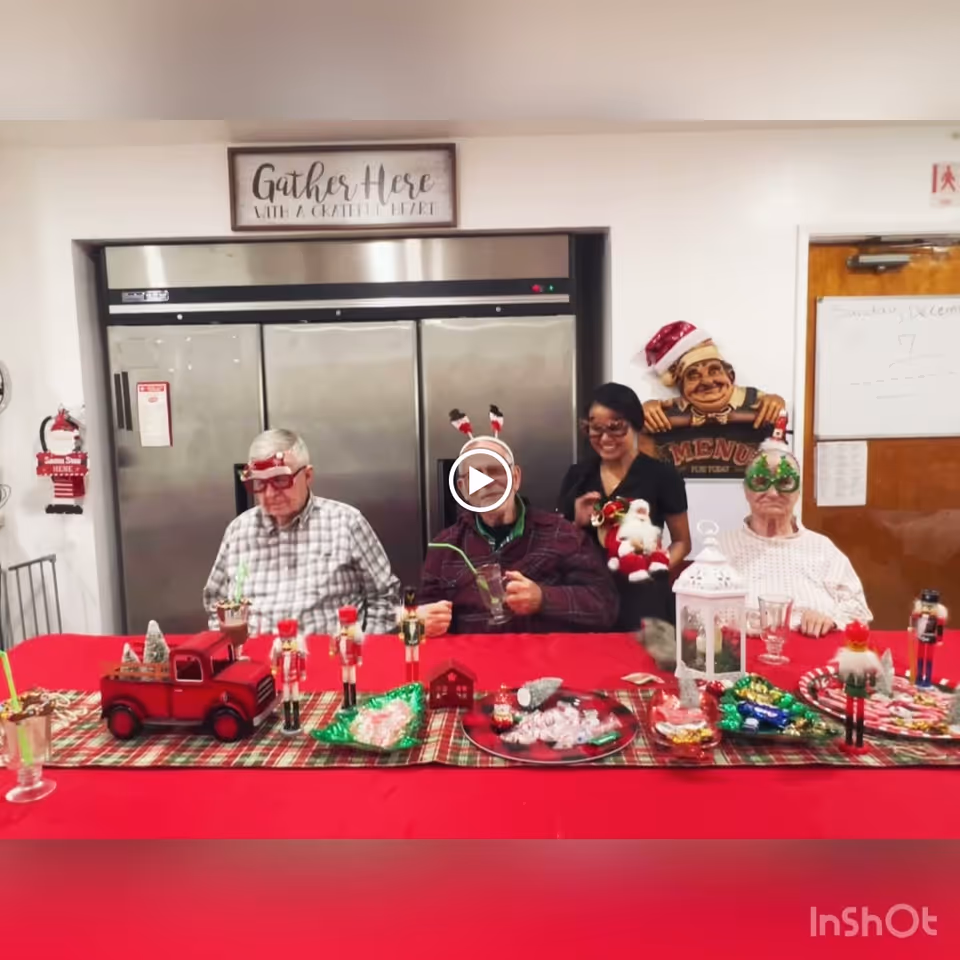 Three elderly individuals and a caregiver sitting at a table decorated with Christmas-themed items including nutcracker figurines, a red toy truck, candy, and a white lantern. The elderly individuals are wearing festive holiday glasses and headbands. Behind them is a large stainless steel refrigerator and a sign that reads 'Gather Here With A Grateful Heart.'