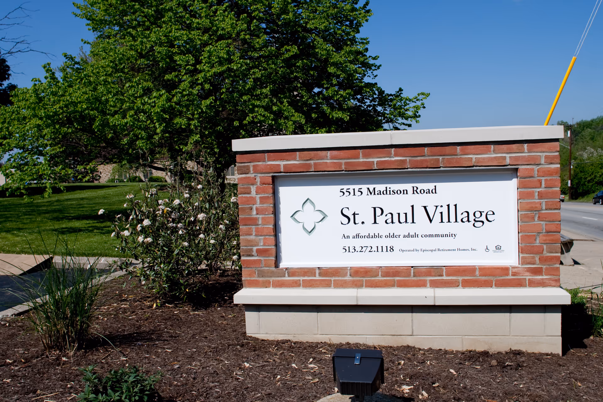A brick sign for St. Paul Village located at 5515 Madison Road, an affordable older adult community, with greenery and a tree in the background under a clear blue sky.