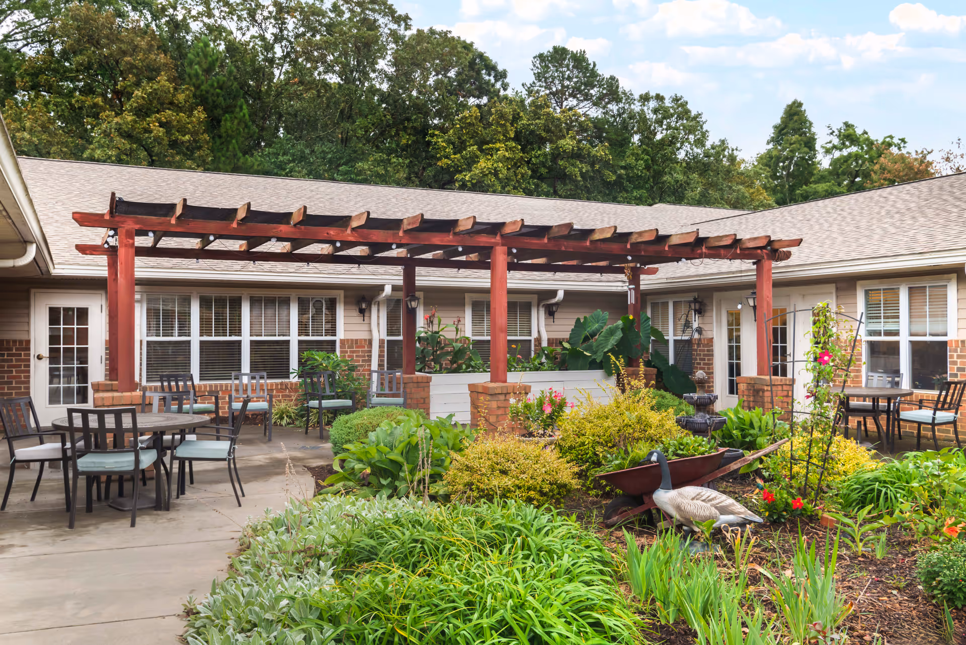 Outdoor patio area at Brookdale Monroe Square featuring a wooden pergola, several tables and chairs, lush green plants, flowers, and a decorative goose statue in a garden bed.